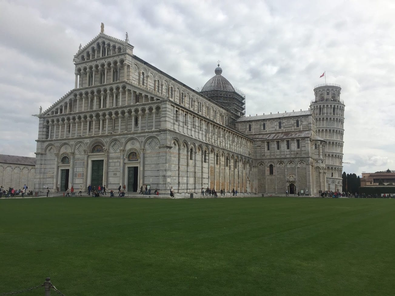 The cathedral and leaning tower of Pisa across the lawn of the Campo dei Miracoli under a flat grey sky