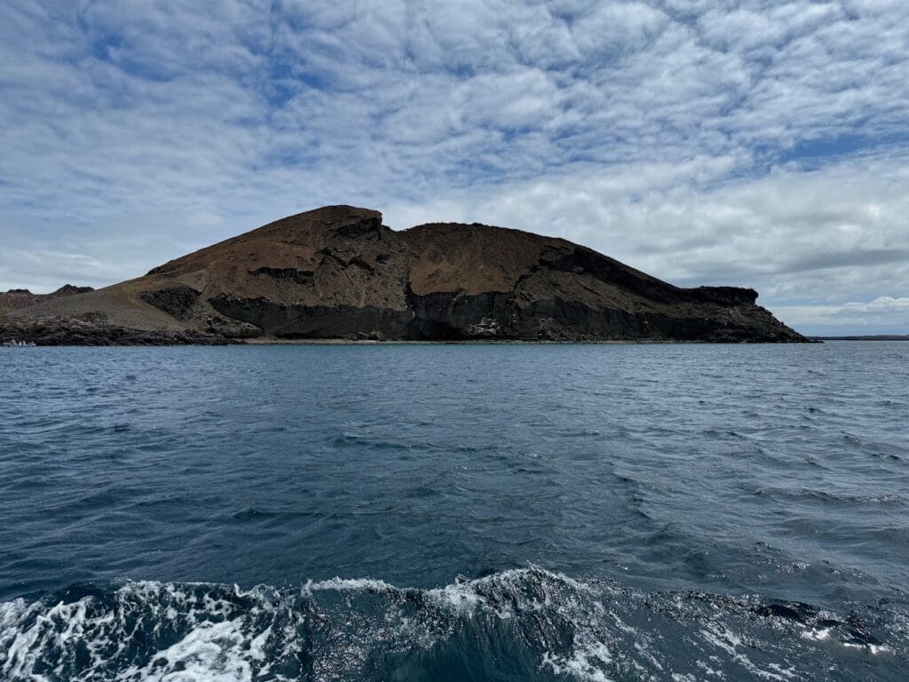 A Galápagos island seen from a boat under a grey cloud deck, with choppy water in the foreground