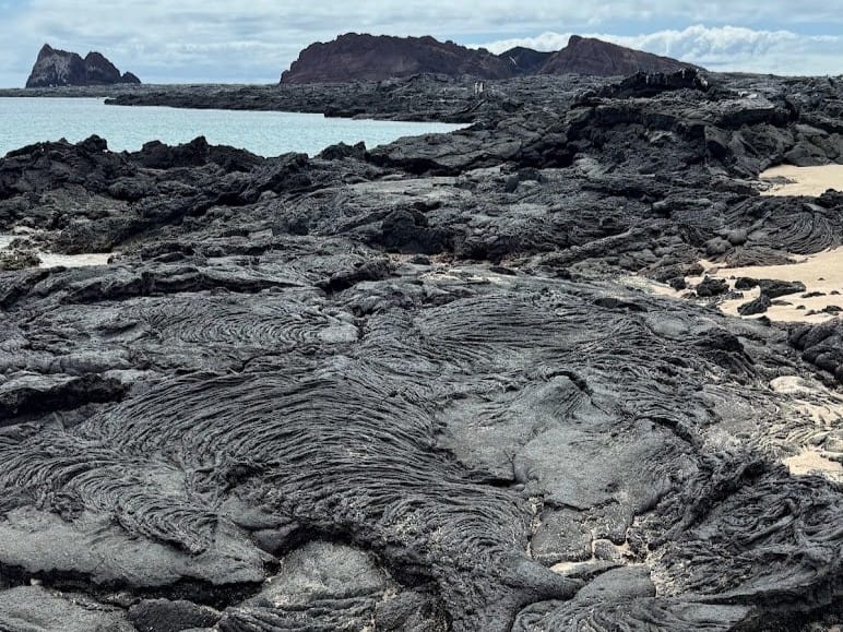 Ropy pahoehoe lava meeting pale sand on a Galápagos shoreline, with footprints across the sand