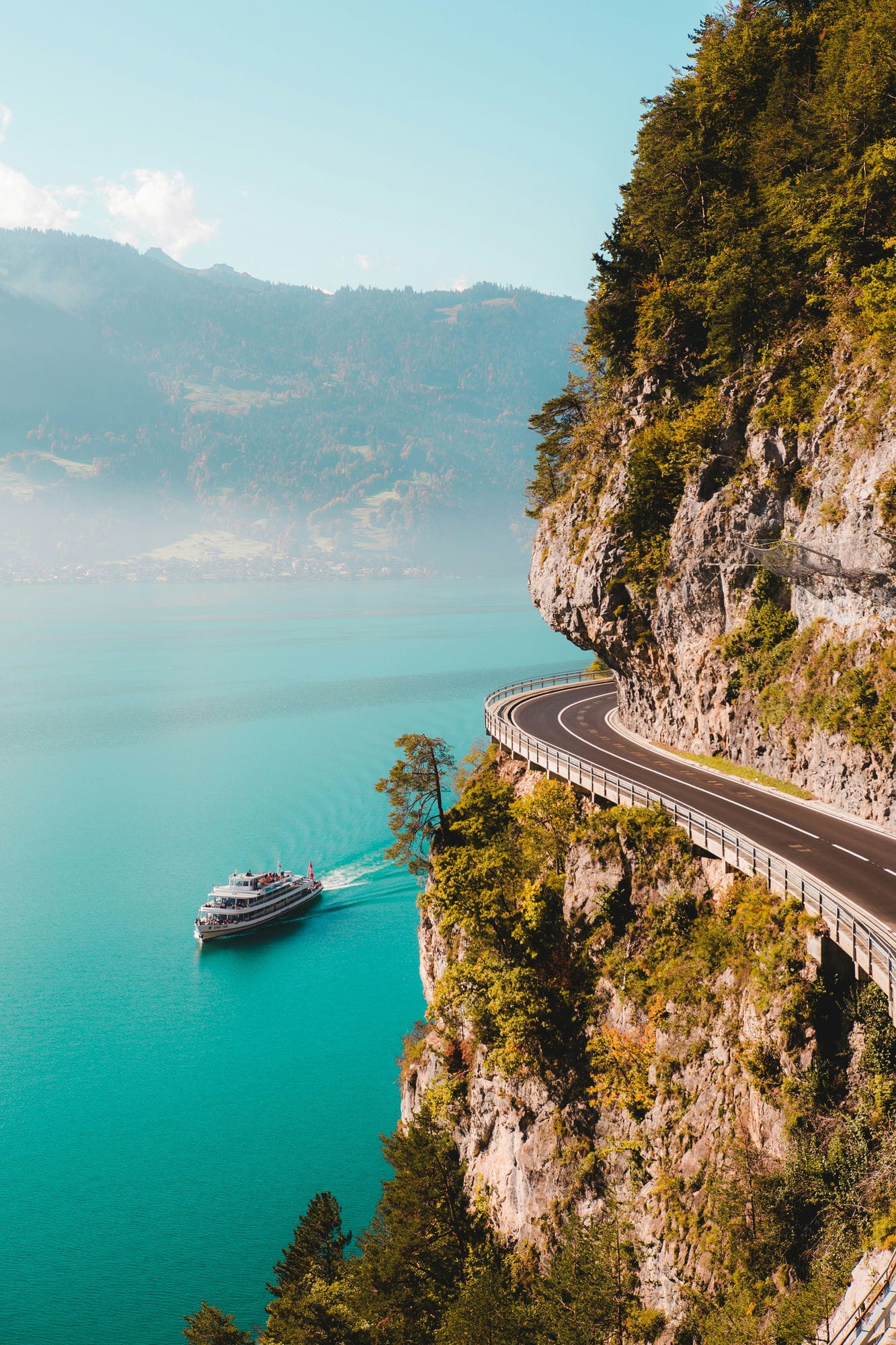 Cruise boat on turquoise water beneath a winding cliffside mountain road in the Alps