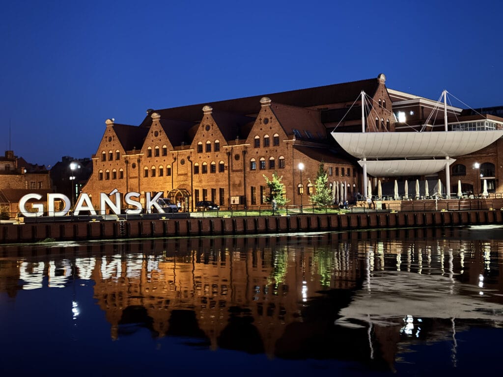 Brick waterfront buildings in Gdańsk lit at dusk and mirrored in the water, with the city's name in tall letters on the quay