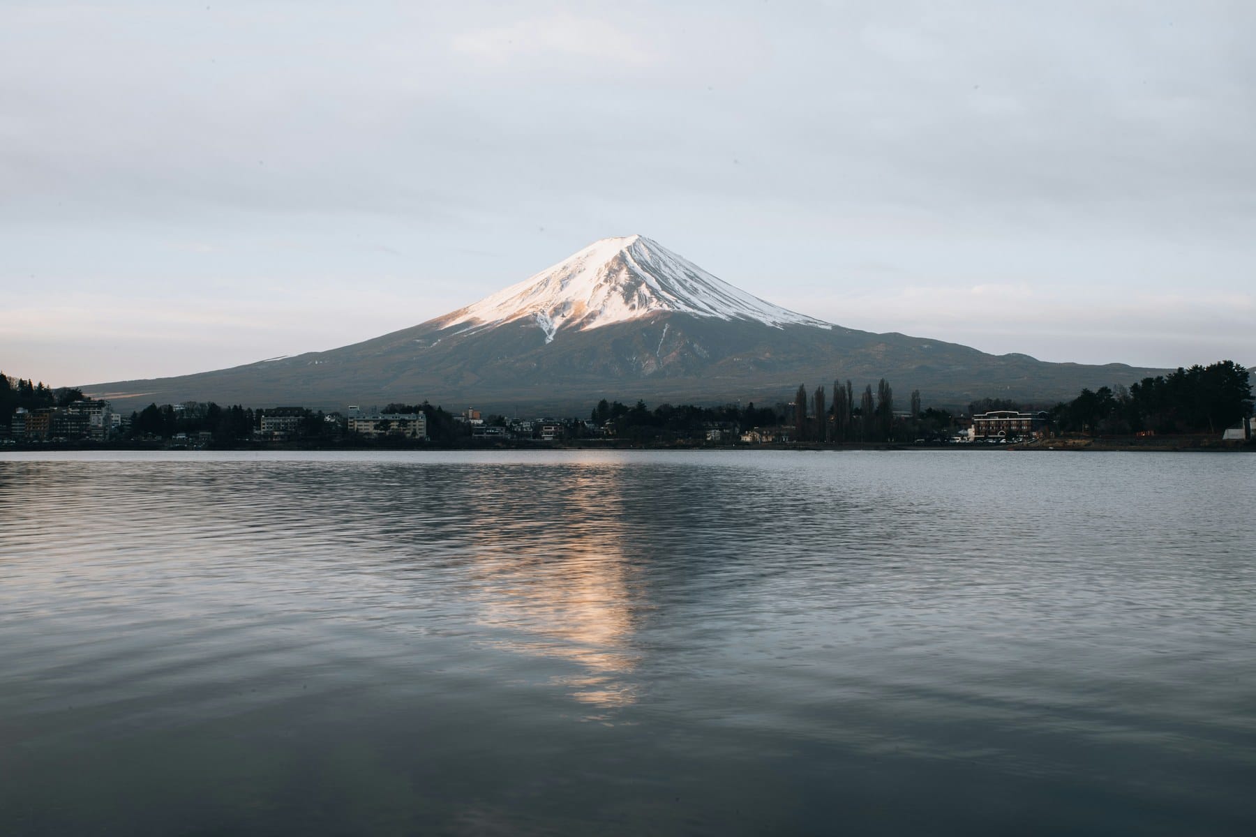 Snow-capped Mount Fuji reflected in a calm lake at dawn, lakeside town in the foreground.