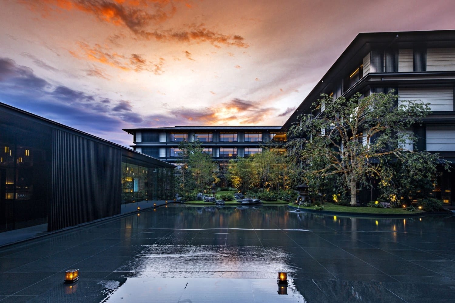 Modern Japanese hotel at sunset with reflecting pond, garden and a large tree, warm lights.
