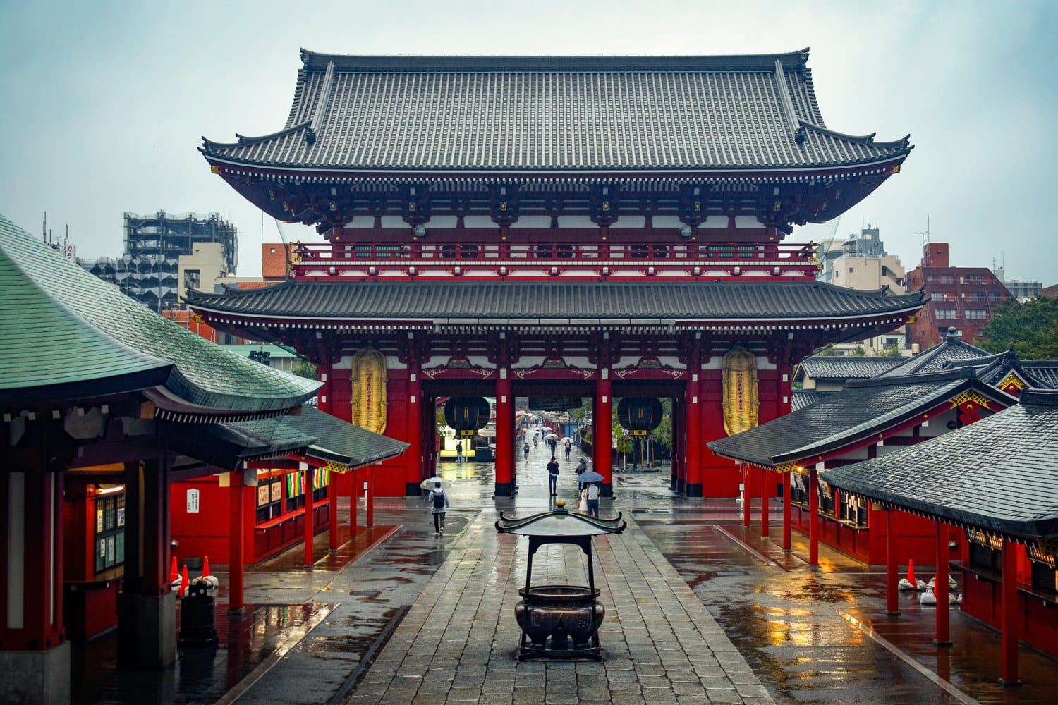 Red two-story temple gate at Senso-ji, Asakusa, wet stone paving, visitors with umbrellas.
