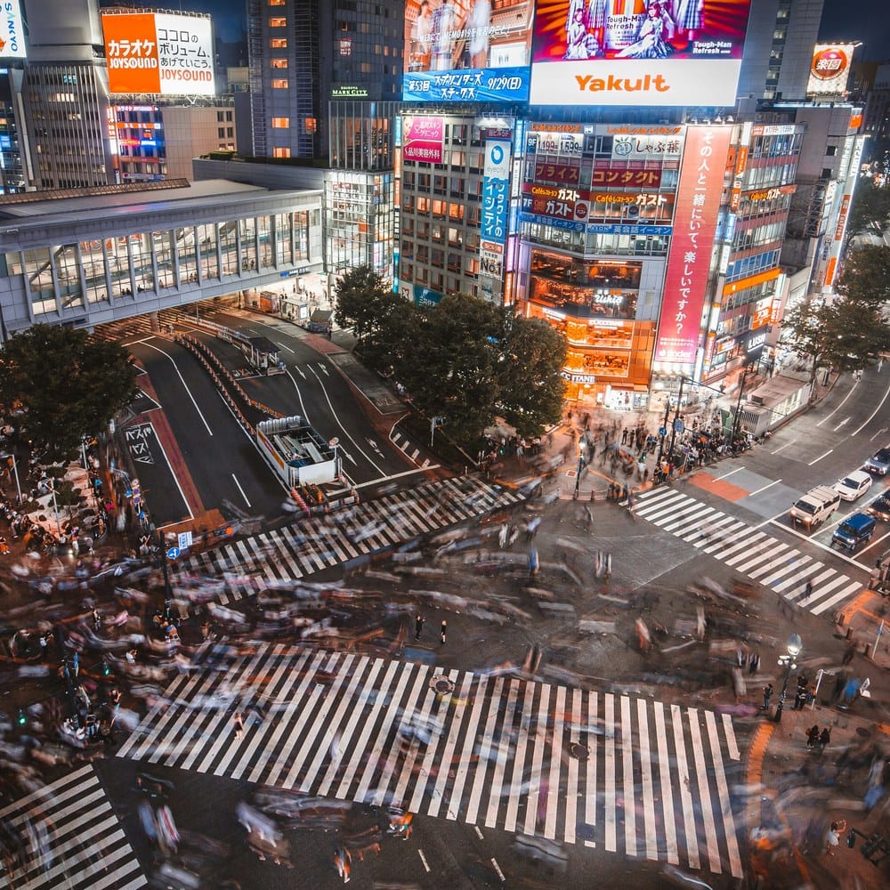 Aerial night view of Shibuya Crossing, blurred crowds and lit billboards.