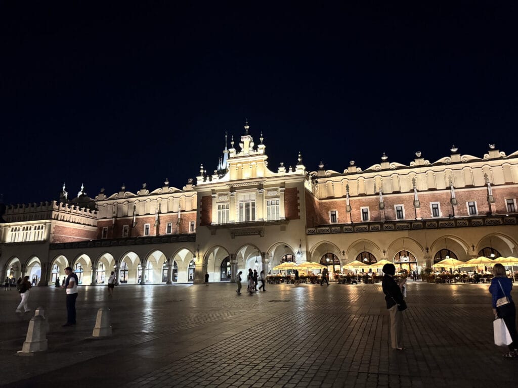 The lit arcades of the Cloth Hall across Kraków's Main Square after dark, café umbrellas glowing under the far end