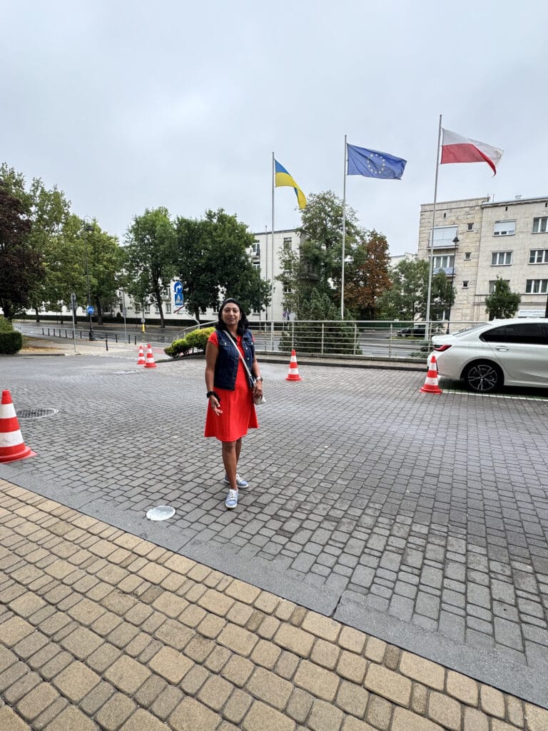 Milli standing outdoors in Warsaw on an overcast morning, Polish and European flags flying behind her