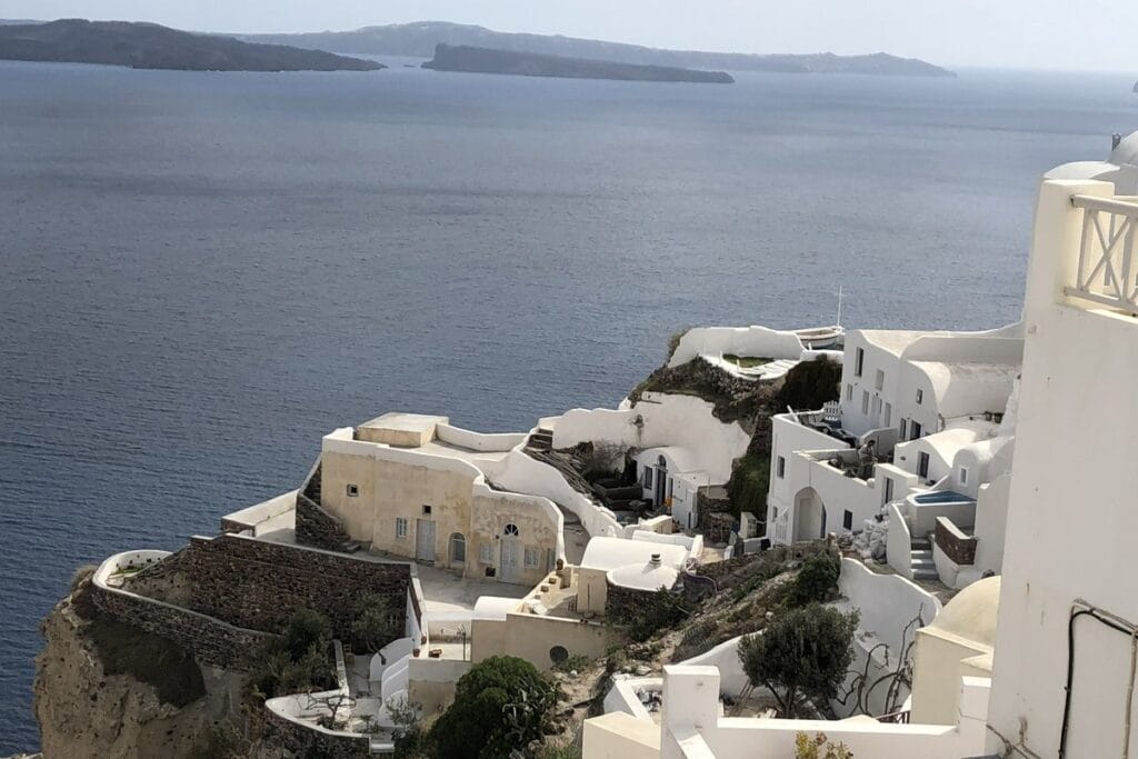 Whitewashed cave houses stepping down the Oia cliff edge above the caldera, with the sea and the far islands beyond