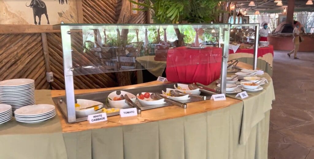 A clean, rustic buffet station at a comfortable East African safari lodge, featuring fresh vegetables like tomatoes and cabbage under a glass sneeze guard.