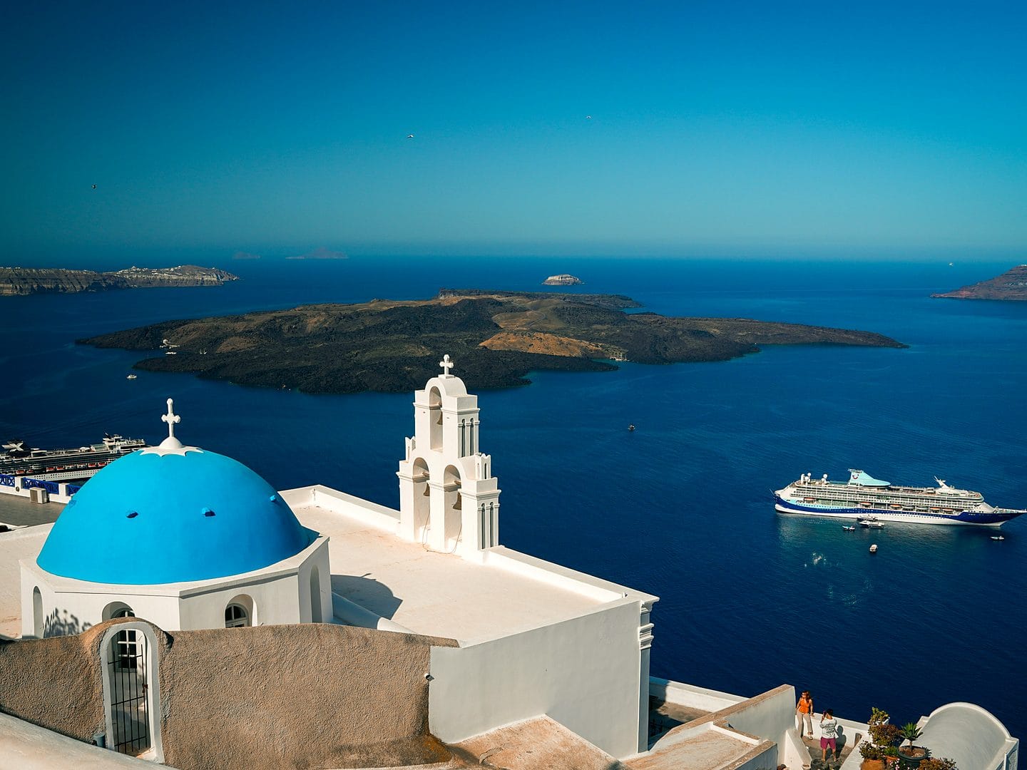 A cruise ship on the deep-blue Aegean Sea seen from Santorini, with the island's white church and blue dome in the foreground