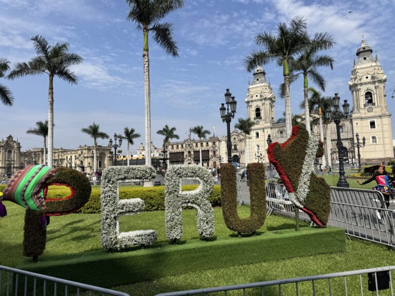 The floral "PERU" letters in Lima's Plaza Mayor, with the twin-towered Lima Cathedral and palm trees behind under a blue sky.
