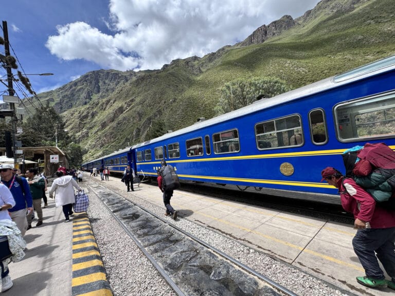 A blue PeruRail train stopped at Ollantaytambo station in the Sacred Valley, green mountains rising behind and travelers on the platform.